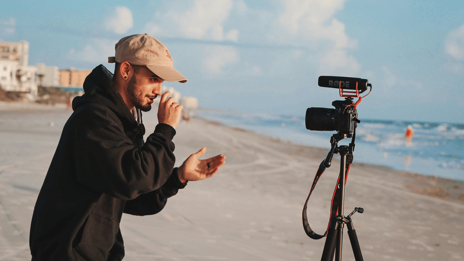  A man records a video of himself on a beach. He will be creating a travel vlog, background music for a vlog will be essential in his process.